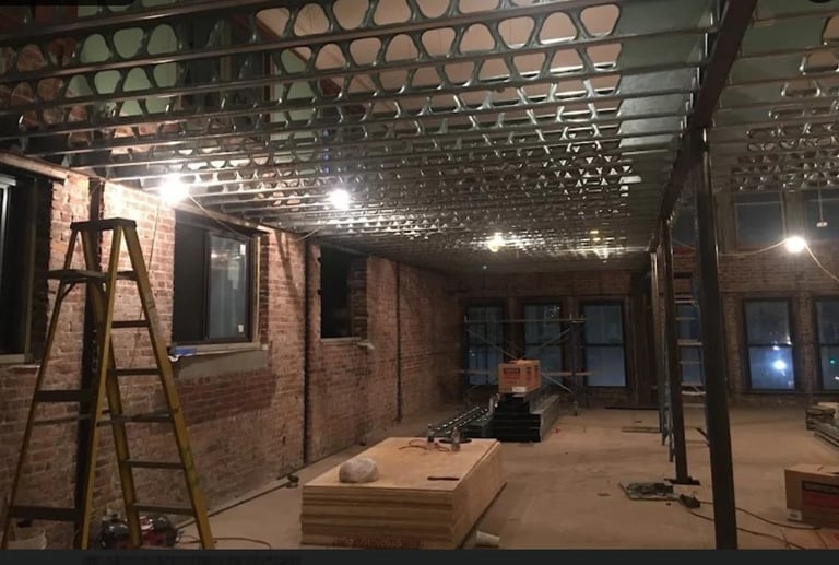 Interior of a brick building under renovation with exposed framework, metal ceiling grid, work materials, and construction lighting