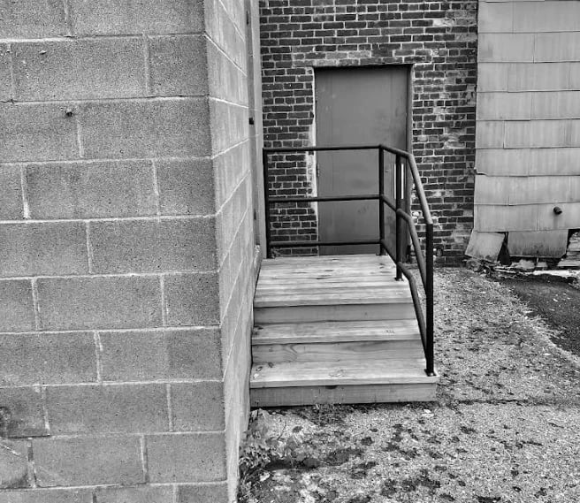 Wooden ramp with metal handrail leading to brick building entrance, black and white photograph