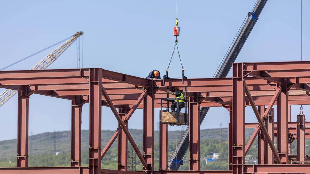 Construction workers operating a crane to lift steel beams during structural assembly of a large industrial building frame.